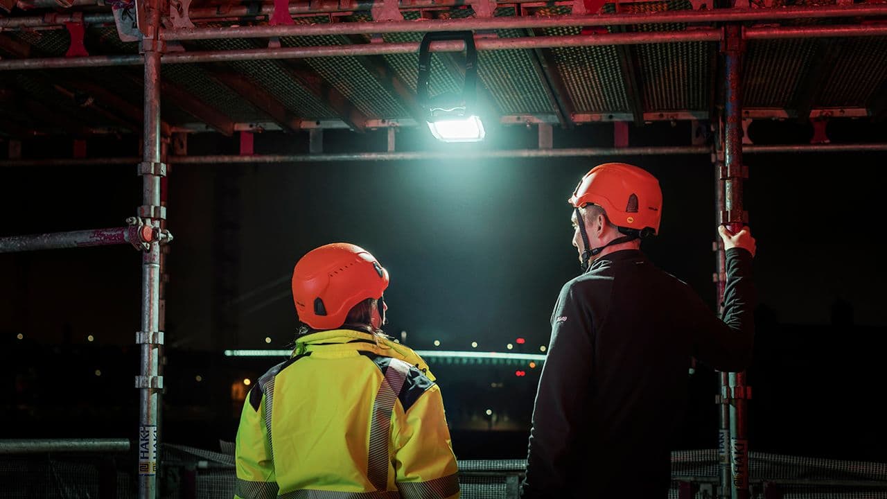 To personer som står på stillas på byggeplass med arbeidslampe festet i stillaset.