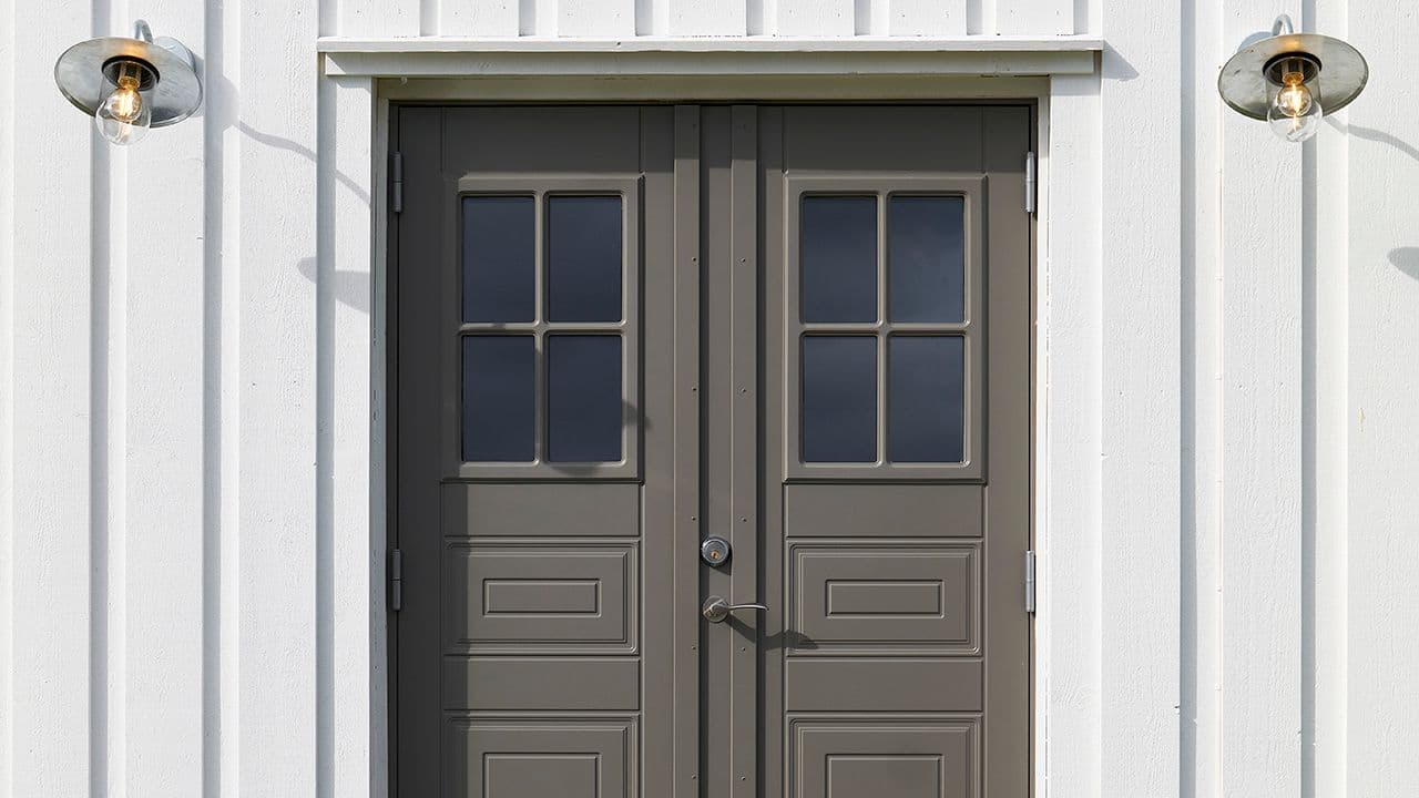 Stående kledning som er hvitmalt med profilerte dobbeltdører og to lamper ved inngangen.