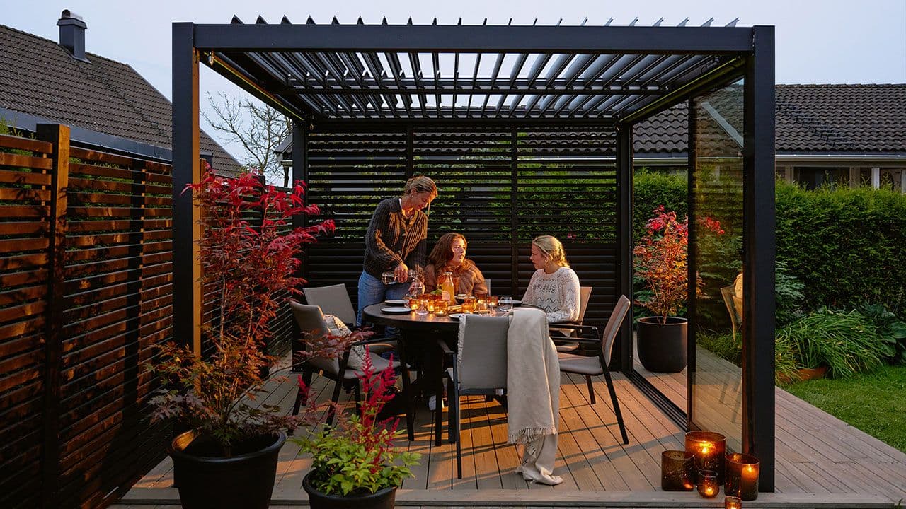 Hagestue aluminium med spisegruppe der tre jenter spiser middag en stemningsfull sommerkveld.