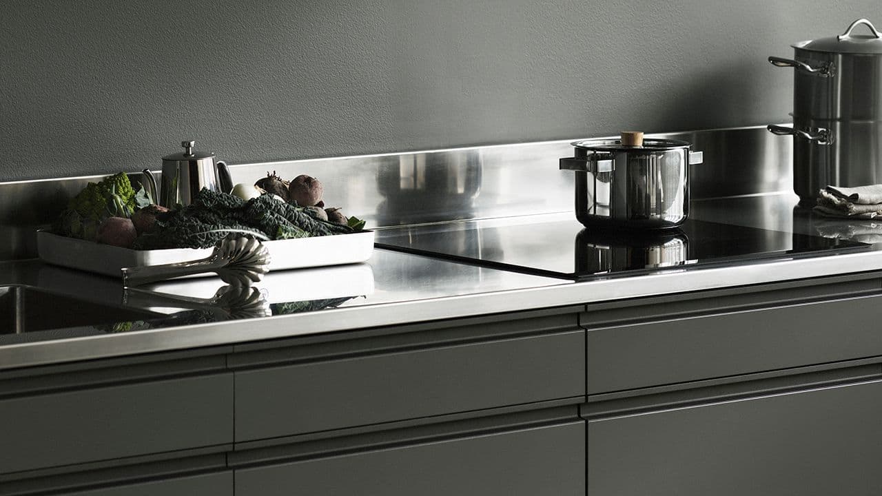 Benkeplate i stål på grønt kjøkken med kasseroller og brett med grønnsaker på kjøkkenbenken.